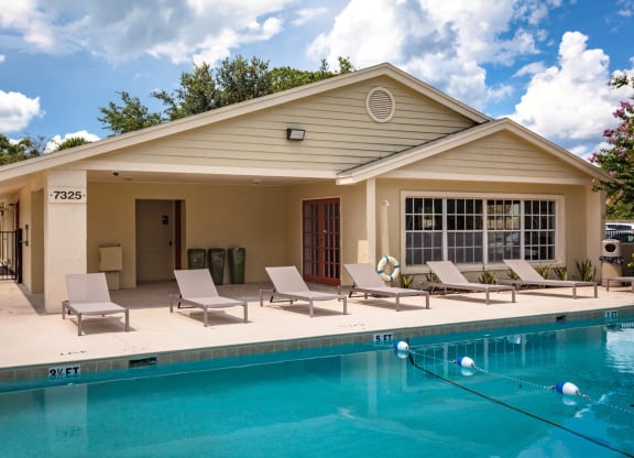 Outdoor swimming pool area at a residential community with lounge chairs, a beige clubhouse building, and a bright blue sky with scattered clouds. The pool features depth markers and a floating safety rope, creating a relaxing recreational space