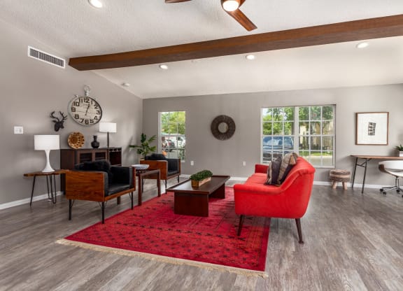 Stylish apartment clubhouse lounge with a red sofa, wooden chairs, and a bold red area rug. The space features modern decor, a vaulted ceiling with wooden beams, large windows, and a cozy workspace with a desk and chair