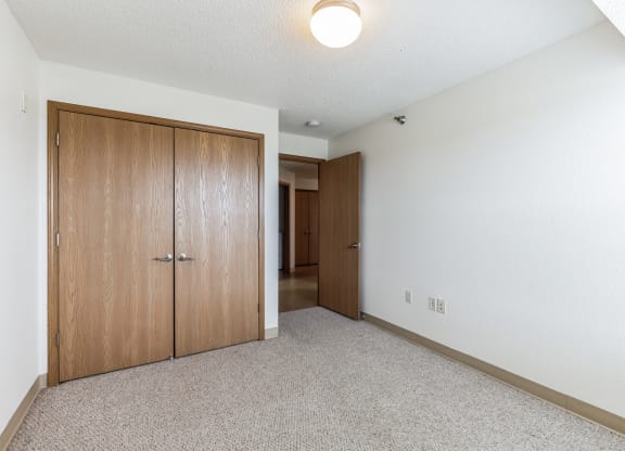 Empty apartment bedroom with white walls, beige carpeting, and a wooden double-door closet. A wooden door is open, leading to a hallway with additional doors visible in the background. The ceiling features a simple round light fixture