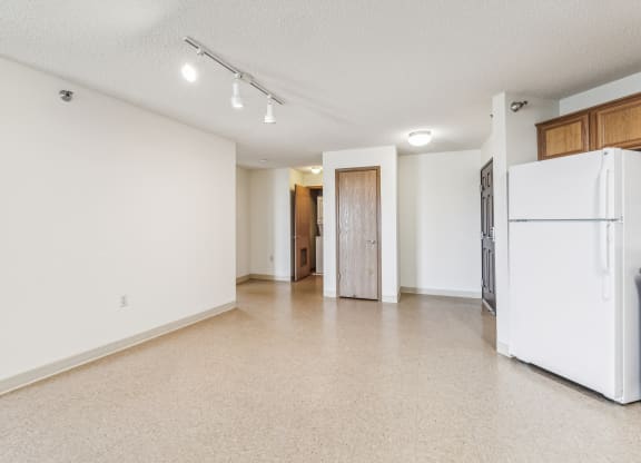 Empty apartment space with beige vinyl flooring, white walls, and track lighting on the ceiling. A white refrigerator is partially visible on the right, with a dark brown entrance door and a closed wooden closet door in the background. An open doorway leads to a laundry area