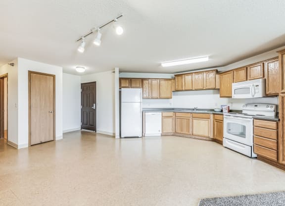 Spacious apartment kitchen with wooden cabinetry, white appliances including a stove, microwave, refrigerator, and dishwasher, and beige flooring. The open layout connects to the living area, with a laundry room and entrance door visible in the background