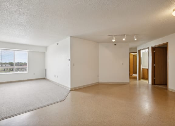 Spacious empty apartment interior with white walls, beige flooring, and a carpeted living area near a large window with natural light. The unit features an open layout with track lighting and multiple doorways leading to bedrooms and a bathroom