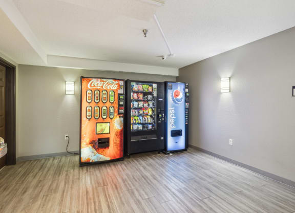 Apartment vending machine area with a Coca-Cola soda machine, a snack vending machine, and a Pepsi soda machine against a gray wall. The space features modern wood-style flooring, soft wall lighting, and a nearby drinking fountain