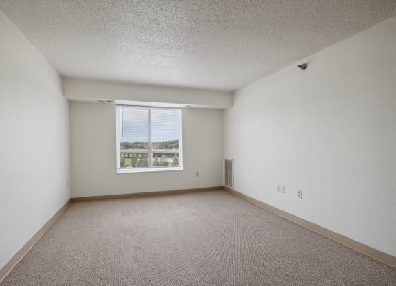 Empty apartment bedroom with white walls, beige carpeting, and a large window with blinds allowing natural light and an outdoor view. Electrical outlets and a vent are visible along the base of the wall
