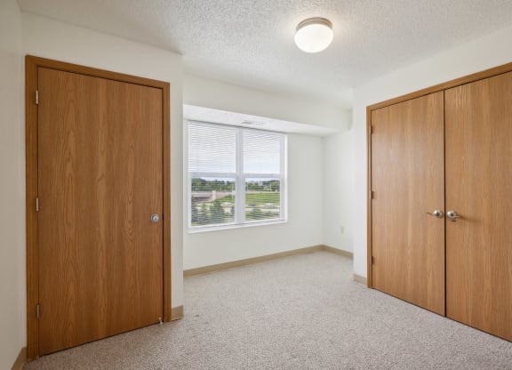 Empty apartment bedroom with white walls, beige carpeting, and a large window with blinds offering a view of green outdoor spaces. The room features two wooden doors—one single door and a double-door closet. A ceiling light fixture is mounted in the center