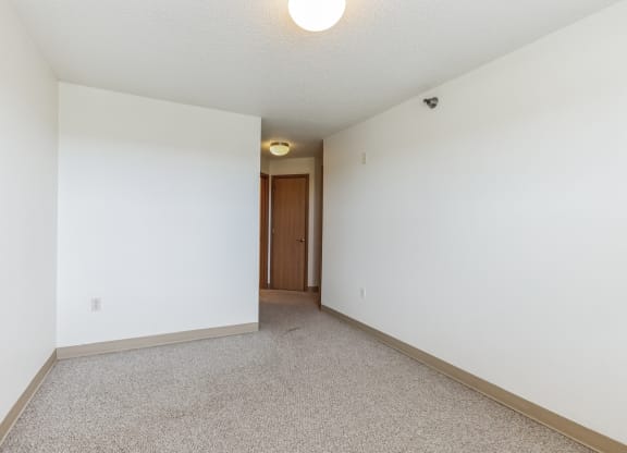 Empty apartment bedroom with white walls, beige carpeting, and a ceiling light fixture. A wooden door in the background leads to a hallway, with another light fixture visible near the entrance