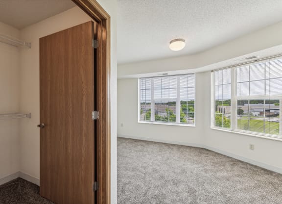 Apartment bedroom with large windows offering a bright view of the outdoors, featuring white walls, gray carpeting, and a ceiling light fixture. An open closet with wire shelving and a wooden door is visible in the foreground