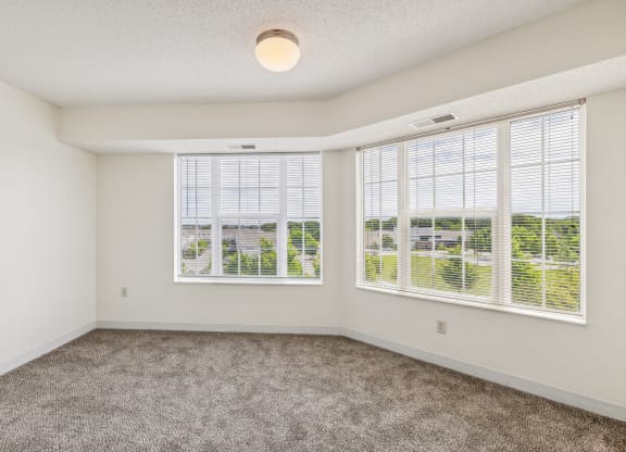 Spacious apartment bedroom with large corner windows featuring white blinds, allowing ample natural light and a scenic outdoor view. The room has white walls, gray carpeting, and a simple ceiling light fixture