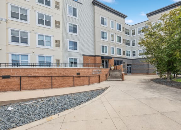 Modern multi-story apartment building with beige and gray siding, large windows, and a landscaped entrance. A sign reading 'Lakewood Apartments' is displayed on a brick retaining wall, with a paved walkway and accessible ramp leading to the main entrance