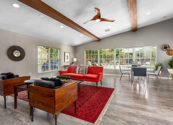 A stylish living room with a red sofa on a vibrant red rug, paired with two dark wood chairs with black fur cushions. Large windows let in natural light, offering a view of the garden. A dining area is seen in the background, and the room features a high ceiling with wooden beams, recessed lighting, and a ceiling fan