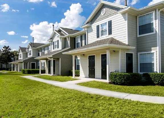 A row of modern townhome-style apartments with beige siding, white trim, and dark front doors, each featuring a small covered porch. The buildings are surrounded by neatly trimmed hedges and a large, well-maintained grassy lawn under a partly cloudy blue sky.