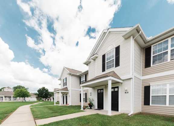 A bright and airy view of townhome-style apartments with beige siding, white trim, and black shutters, featuring dark front doors and small covered porches. The homes are surrounded by green lawns and a concrete walkway, set against a partly cloudy blue sky.