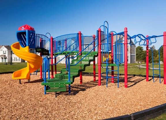 A colorful playground set featuring blue, red, green, and yellow components, including slides, climbing structures, and monkey bars. The playground is surrounded by wood chips and open grassy areas, with residential buildings visible in the background under a clear blue sky.
