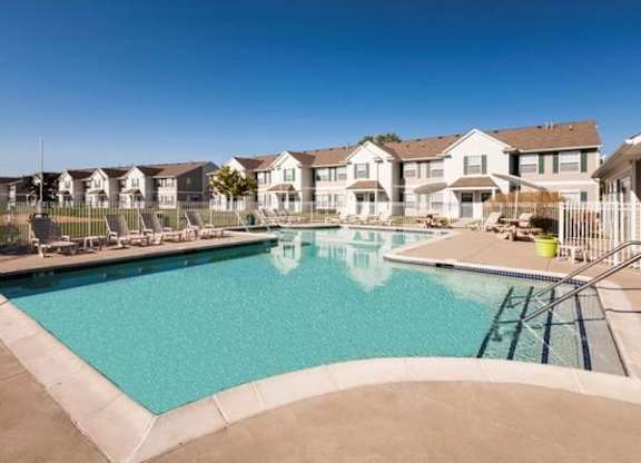 A sparkling outdoor swimming pool surrounded by a concrete deck with lounge chairs and umbrellas. The pool area is adjacent to a row of townhome-style apartments with beige siding and white trim, set against a clear blue sky.