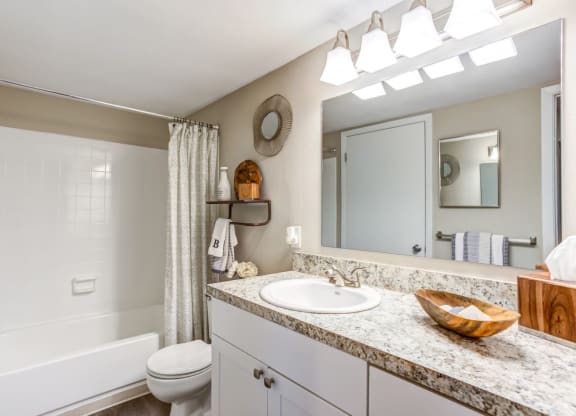A bathroom featuring a white bathtub with a gray and white shower curtain, a white sink with a granite countertop, and a large mirror. A small shelf holds decorative items and towels, and a wooden bowl adds a natural touch to the countertop