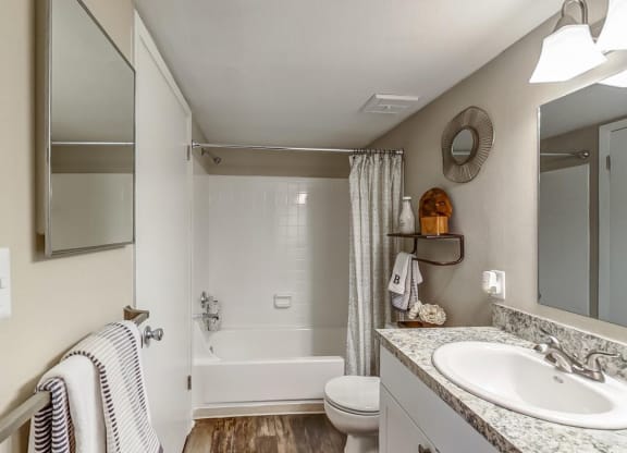 A bathroom with a white bathtub and shower, a granite countertop with a white sink, and a large mirror above the sink. Towels are neatly arranged on the towel bar, and a small wooden shelf holds decorative items and a towel