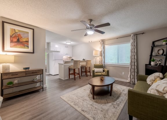 Modern open-concept living room and kitchen with stylish decor, featuring green sofas, a round wooden coffee table, a ceiling fan, and a breakfast bar with beige stools. The space is well-lit with natural light from a large window with patterned curtains and warm ambient lighting