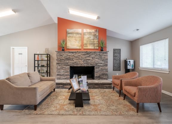 Cozy clubhouse lounge with a stone fireplace, beige sofa, two orange accent chairs, and a modern coffee table on a patterned area rug. The space features warm lighting, an orange accent wall with abstract artwork, and decorative plants