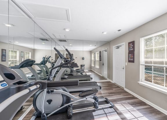 Modern fitness center with treadmills, ellipticals, and exercise bikes lined up against a mirrored wall. The gym has large windows allowing natural light, light wood flooring, and motivational wall art. A door leads to an outdoor area with a view of a pool through the window, creating a bright and inviting workout space