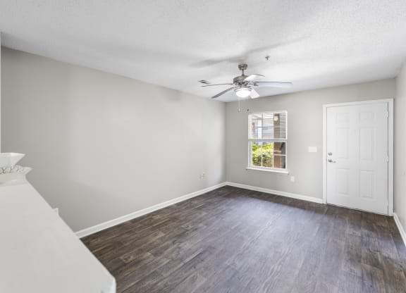 Modern apartment living area with dark wood-style flooring, light gray walls, and a ceiling fan with a built-in light fixture. A white entry door and a window allowing natural light are visible, with a partial view of a countertop in the foreground
