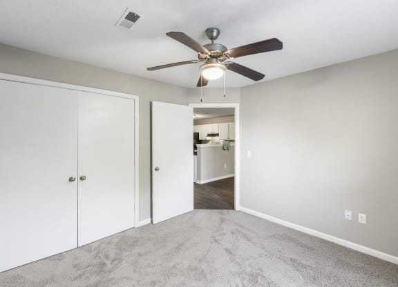 Modern apartment bedroom with light gray walls, plush gray carpeting, and a ceiling fan with a built-in light fixture. A white double-door closet is on one side, and an open door leads to a kitchen with white cabinetry and dark wood-style flooring
