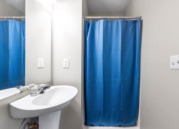 Compact apartment bathroom featuring a white pedestal sink with a mirror above, light gray walls, and a shower with a blue curtain. Electrical switches and outlets are visible on the wall, adding functionality to the space