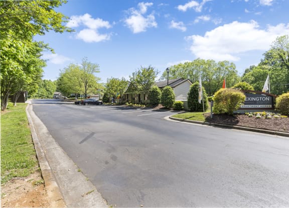 Entrance to Lexington Apartment Homes with a well-maintained landscape, a prominent community sign, and neatly trimmed bushes. The paved road leads into the complex, surrounded by lush greenery and trees under a bright blue sky with scattered clouds. A few cars are parked near the leasing office, creating a welcoming residential atmosphere