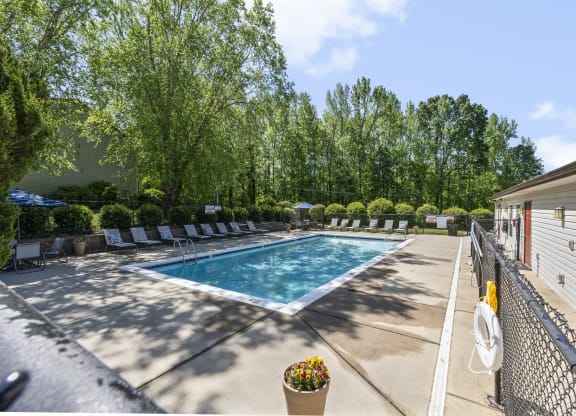 Outdoor swimming pool surrounded by lounge chairs and lush greenery. The pool is enclosed by a fence with a pool house or clubhouse on the right side. A bright blue sky and tall trees provide a serene backdrop, with shadows cast on the concrete deck. A potted flower plant sits near the pool entrance, adding a touch of color to the well-maintained space