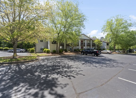 Apartment complex with spacious parking lot surrounded by lush green trees. The residential buildings have balconies and well-maintained landscaping, creating a welcoming environment. A few cars and a pickup truck are parked, while shadows from trees cast across the asphalt under a clear blue sky