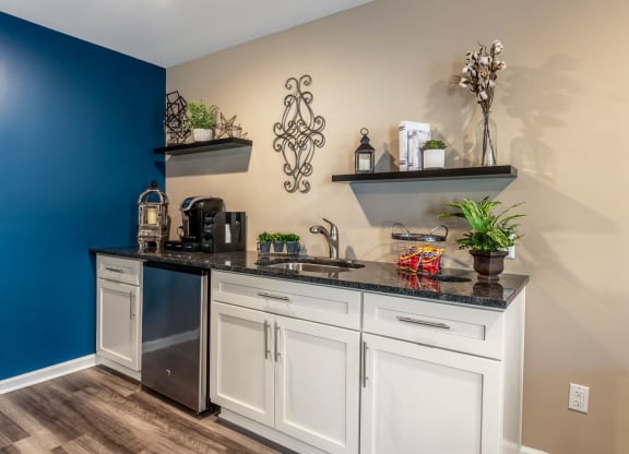 Stylish kitchenette with white cabinets, black countertops, and a small stainless steel fridge. Floating shelves with plants and decor complement the beige and blue walls