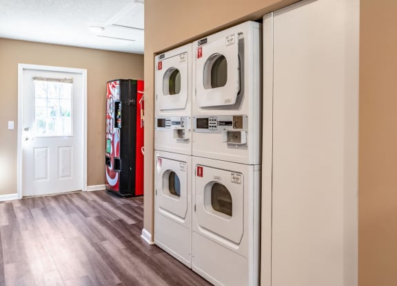 Apartment community laundry room featuring stacked white commercial dryers against a beige wall. The space has wood-style flooring, bright lighting, and a vending machine near the entrance door with natural light coming through a windowed panel