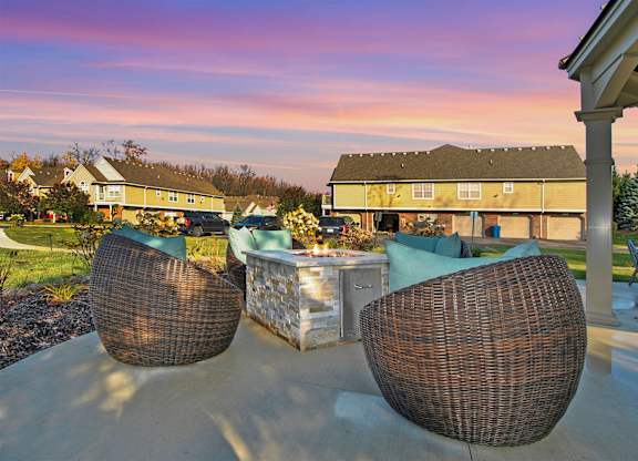 Two wicker chairs are on a patio with a table in front of a house.
