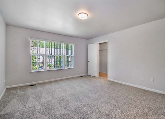 A room with a carpeted floor, a window with a view of a fence and trees, and a door leading to another room.