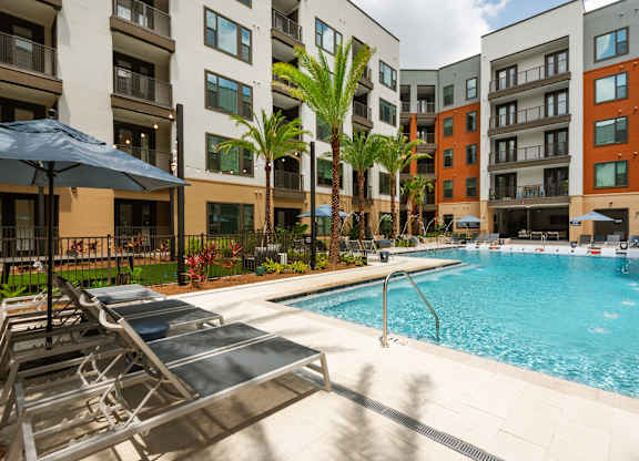 A pool surrounded by lounge chairs and umbrellas in front of apartment buildings.