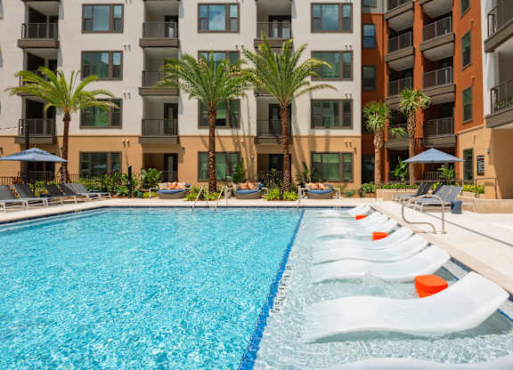 A swimming pool with lounge chairs and palm trees in front of a multi-story apartment building.