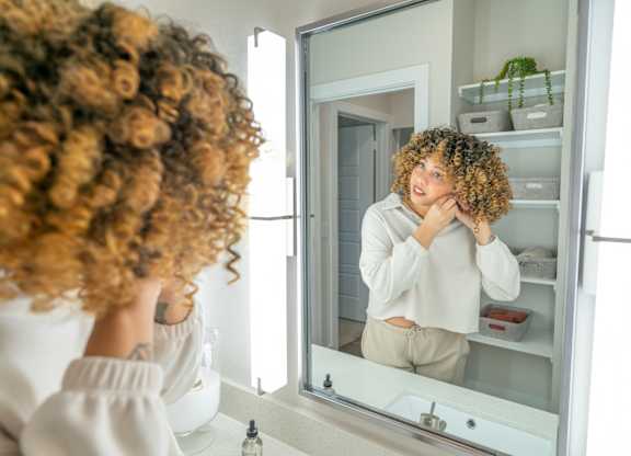 A woman with curly hair is looking at her reflection in a bathroom mirror.