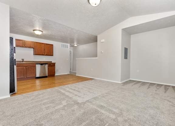 A spacious room with a carpeted floor and a kitchen area with wooden cabinets and a stainless steel dishwasher.