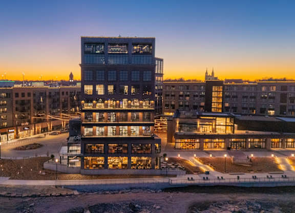 A modern building with a glass facade is illuminated from within, set against a twilight sky.
