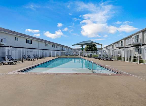 A swimming pool in a sunny day surrounded by chairs and buildings.