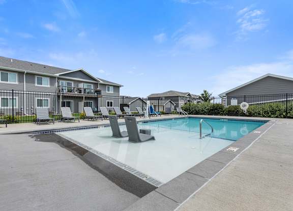 A swimming pool surrounded by a concrete patio and lounge chairs in front of apartment buildings.