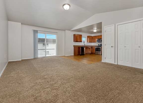 A spacious living room with a carpeted floor and a kitchen area in the background.