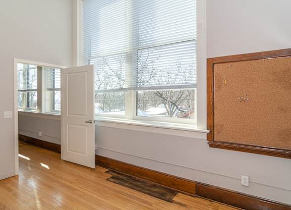 A room with a white door, a window with blinds, and a corkboard on the wall.