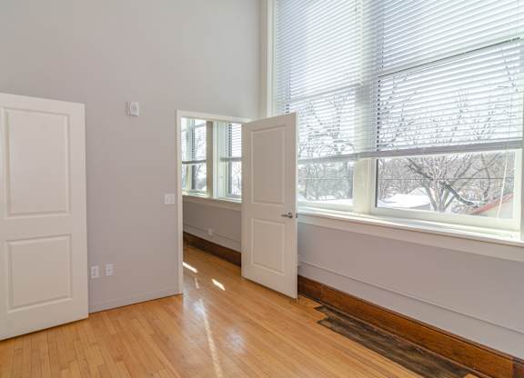 A room with white walls and wooden floors has two white doors and a window with blinds.