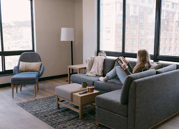 A woman is sitting on a grey couch in a living room with a large window behind her.