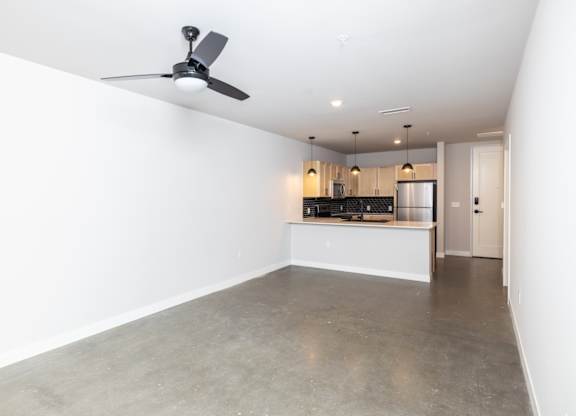 A room with a ceiling fan and a kitchen area in the background.