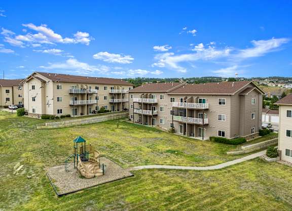 A playground with a slide is in the middle of a grassy area in front of apartment buildings.