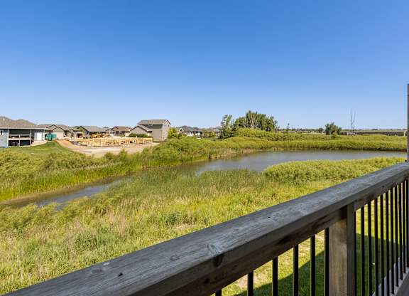 A wooden deck overlooks a grassy field with a river and houses in the distance.