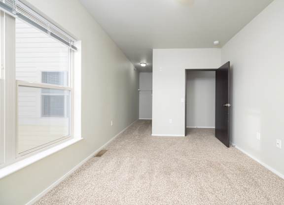 A room with a carpeted floor, a window with blinds, and a door leading to another room.