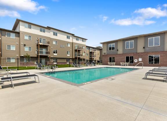 A swimming pool is surrounded by sun loungers in front of apartment buildings.
