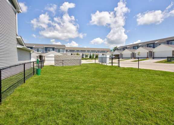 A grassy area in front of a row of houses with a fence and a green trash can.